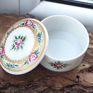 May include: A small, white porcelain trinket box with a floral design. The lid features a gold rim and a central floral motif with pink, red, and green roses. The box is open, revealing a white interior.