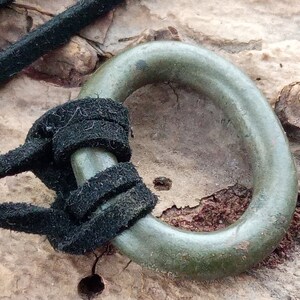Op de afbeelding: Een groene metalen ring met een zwart koord erdoorheen geregen. De ring is met een knoop aan het koord vastgemaakt.