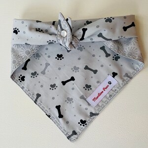 May include: A gray dog bandana with black paw prints and bone designs. The bandana has a white label that says "Maritime Paws".
