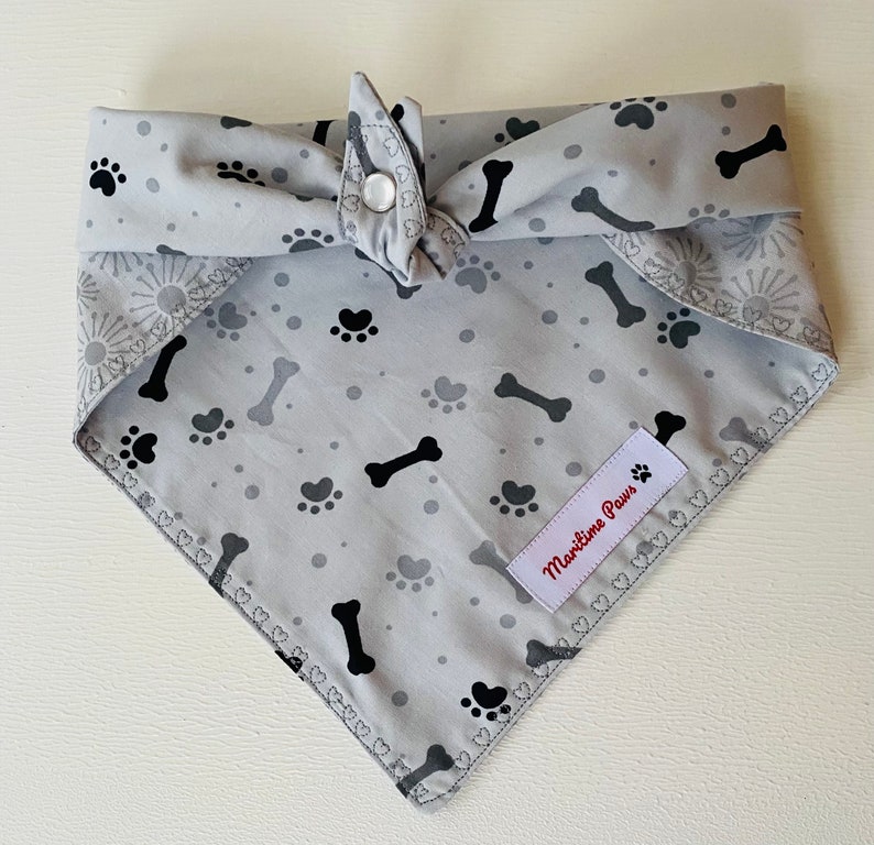 May include: A gray dog bandana with black paw prints and bone designs. The bandana has a white label that says "Maritime Paws".