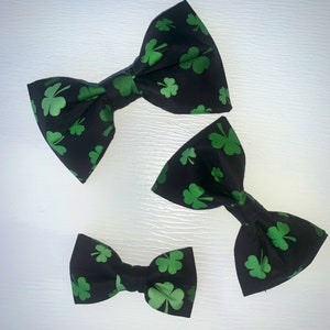 May include: Three black bow ties with green shamrock patterns. The bow ties are different sizes, with the largest bow tie at the top of the image and the smallest bow tie at the bottom.