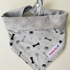 May include: A gray dog bandana with a black paw print and bone pattern. The bandana has a white button closure and a red label that says "Maritime Paws".