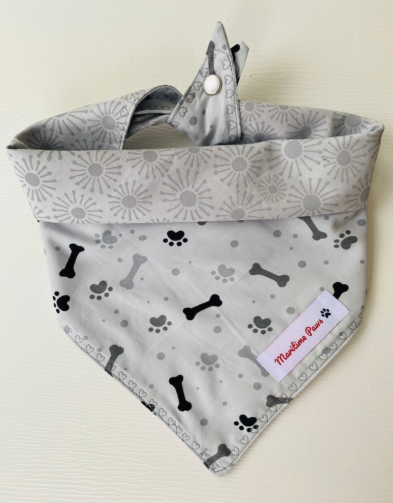 May include: A gray dog bandana with a black paw print and bone pattern. The bandana has a white button closure and a red label that says "Maritime Paws".