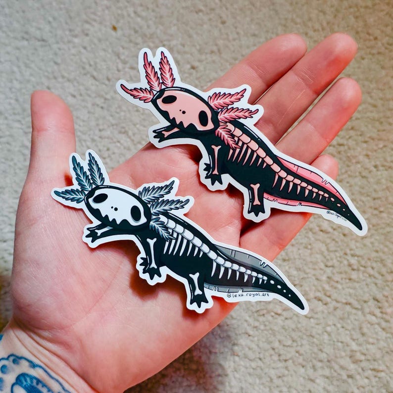 May include: Two axolotl skeleton stickers with black and white skeletons and pink and black details. The stickers are on a hand.