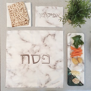 May include: A white marble Passover Seder plate with the Hebrew word "Pesach" in brown lettering. The plate is set with a small plate of matzah, a small plate of carrots, potatoes, and parsley, and a small plate of cheese and a rolled piece of dough.