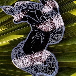 May include: An embroidered patch featuring a black and white design of two hands intertwined in a graceful, flowing motion. The intricate stitching creates a high-contrast graphic effect with a light-colored border outlining the shape of the hands.