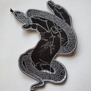 May include: Black and gray embroidered iron-on patch of a hand holding a snake. The snake is wrapped around the hand and its tail is curled up.