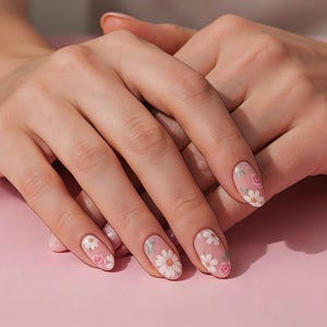 May include: Oval-shaped fingernails featuring a clear base coat adorned with delicate white daisy and pink rose floral nail art designs. The manicure displays a clean, rounded edge finish on each nail, showcasing a detailed botanical pattern.