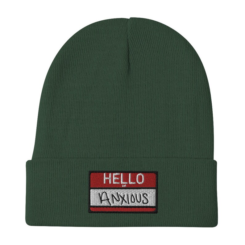 May include: A dark green beanie with a red and white name tag that says "HELLO ANXIOUS" in white lettering.