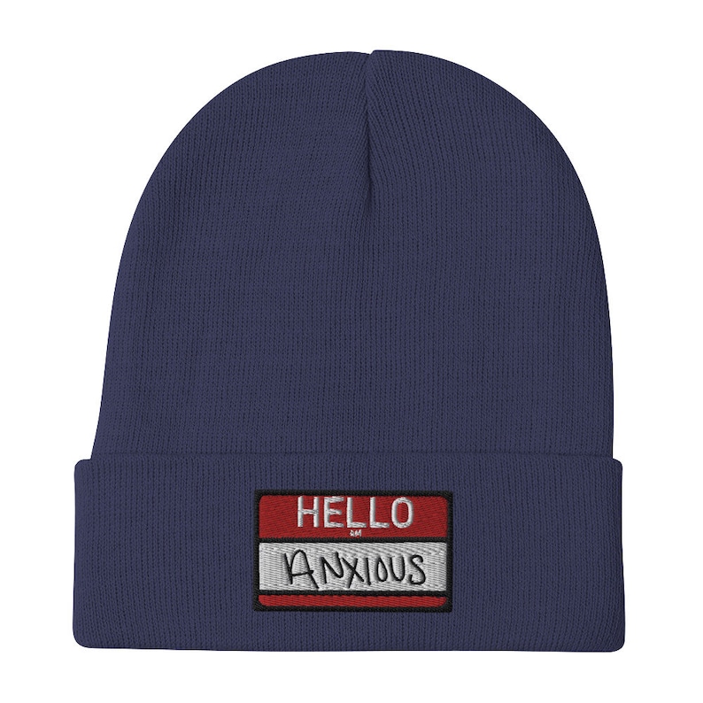 May include: A navy blue beanie with a red and white name tag that says "HELLO ANXIOUS" in white embroidery.