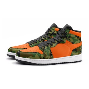May include: High-top sneakers with a vibrant orange and camouflage design. The shoes feature a black tongue, laces, and collar, with a white sole. The camouflage pattern incorporates shades of green, brown, and black.