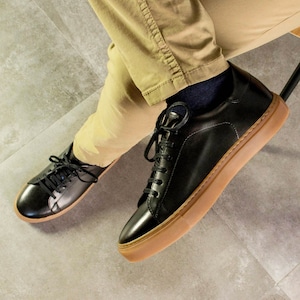 May include: Black leather sneakers with brown soles and black laces. The shoes are shown from a low angle, with the laces neatly tied. The sneakers are paired with khaki pants and dark blue socks, suggesting a casual yet stylish look.