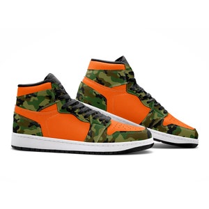 May include: High-top sneakers in orange and camouflage green. The shoes feature a white sole, black laces, and a black collar. The design incorporates a vibrant orange color on the side panels and a camouflage pattern on the overlays.