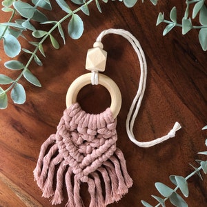 May include: A handmade macramé car charm in a dusty rose colour. The charm features a wooden ring and a geometric wooden bead. The macramé design has a woven pattern with fringe. The charm hangs from a natural-coloured cord.