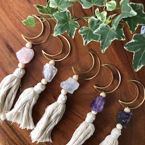 Könnte beinhalten: Sechs goldene Halbmond-Anhänger mit Kristallsteinen und weißen Quasten. Die Kristalle sind rosa, klar, hellblau, lila und dunkelviolett.