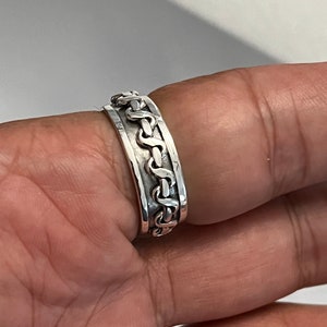 May include: A silver ring with a braided design. The ring is worn on a finger.