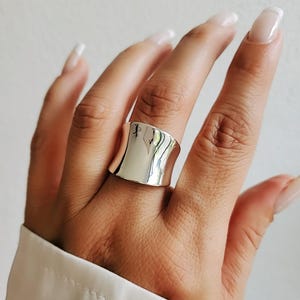 Puede incluir: Un anillo de plata con una banda ancha y curva. El anillo es liso y brillante.