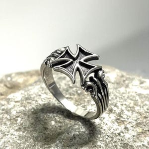 Könnte beinhalten: Ein silberfarbener Metallring mit einem markanten Eisernen Kreuz als Mittelstück, das eine dunkle, vertiefte Oberfläche aufweist. Die Ringschiene zeigt komplexe, erhabene lineare Details, die vom Kreuz ausgehen und eine Textur erzeugen.
