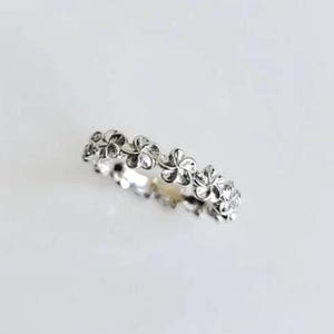 Può includere: Un anello color argento caratterizzato da una fascia continua di piccoli fiori a cinque petali collegati tra loro. Il metallo ha una finitura lucida e riflettente che cattura la luce, creando un motivo floreale ripetuto lungo tutta la circonferenza.