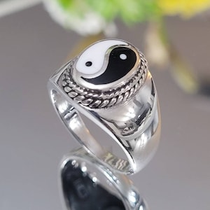May include: A silver ring featuring a black and white yin yang symbol. The oval-shaped symbol is set in a textured, rope-like border. The ring has a polished, reflective surface.