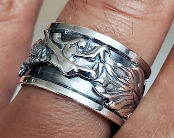 Può includere: Un anello d'argento con un design dettagliato raffigurante una figura mitica ed elementi floreali. L'anello è indossato su un dito, mettendo in risalto l'intricata lavorazione artigianale e la finitura lucida. L'anello ha una fascia rotante.