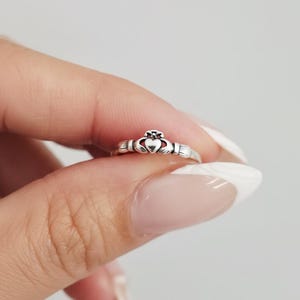 May include: A silver Claddagh ring featuring a heart, crown, and hands design. The ring is held between fingers, showcasing its intricate details. The Claddagh ring is a traditional Irish ring representing love, loyalty, and friendship.