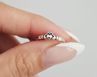 Può includere: Un anello Claddagh in argento, un design tradizionale irlandese, con un cuore, una corona e mani. L'anello è tenuto tra le dita, evidenziando i suoi dettagli. L'anello Claddagh simboleggia amore, lealtà e amicizia.