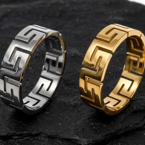 May include: Two rings with a Greek key design. One is silver, the other gold. They sit on a dark, textured surface. The rings feature a repeating geometric pattern, a classic design element.