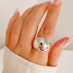 May include: A large, polished silver ring with a rounded, smooth surface. The ring is worn on a finger, reflecting the surrounding environment. The ring's design is simple and modern, suitable for everyday wear.