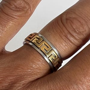 May include: A two-tone ring with a silver band and a gold-colored Greek key design. The ring is worn on a finger, showcasing the intricate pattern and contrasting colors. The ring's design is a classic and elegant style.