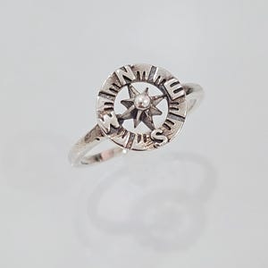May include: A silver ring with a compass design. The compass features a star with a small pearl in the center and the letters N, E, S, and W around the edge.