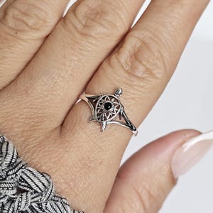 May include: A silver ring featuring a turtle design with a black gemstone center. The turtle's shell has an openwork design. The ring is worn on a finger, showcasing its intricate details and craftsmanship. The ring's band is thin and delicate.