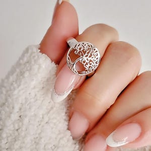 May include: A silver ring with a tree of life design. The ring is on a finger with a white and pink French manicure.