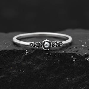 Peut inclure: Une bague en argent avec une perle centrale et des accents perlés décoratifs. La bague est posée sur un fond sombre et texturé, soulignant son design délicat et son savoir-faire. Un accessoire de bijouterie classique et élégant.