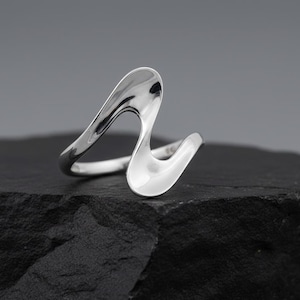 May include: A polished silver ring with a flowing, abstract design. The ring features a wave-like shape with smooth, reflective surfaces. The ring is displayed on a dark, textured surface.