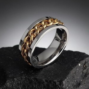 May include: A silver ring with a gold chain design. The ring's central chain is gold, set between silver bands. The ring is displayed on a dark, textured surface, creating a contrast.