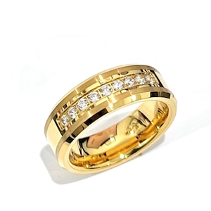 Puede incluir: Un anillo dorado con una fila de piedras claras y brillantes. El anillo tiene una banda ancha con un acabado pulido y un engaste en canal para las piedras. El anillo se presenta sobre un fondo blanco.