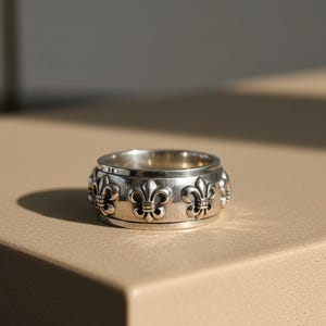May include: A silver ring featuring a repeating fleur-de-lis design. The ring is broad and has a textured finish. It is presented on a neutral surface, with a shadow cast by sunlight.