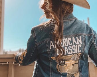 May include: A blue denim jacket featuring a large graphic on the back with the text "AMERICAN CLASSIC" in white block letters above an illustration of a vintage airplane and the text "Since 1938" in white script, worn with a tan cowboy hat.