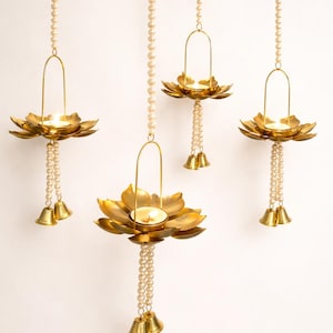 May include: Four hanging candle holders with a lotus flower design. The gold-colored holders are suspended by strings of pearls. Each holder has a small candle inside and is adorned with bells at the bottom. The background is plain white.