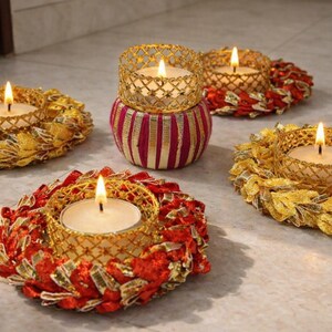 May include: Candle holders with lit candles. The holders have gold and red detailing, with a gold filigree design around the candles. The candles are on a light surface, creating a warm atmosphere. The candles are a decorative item.