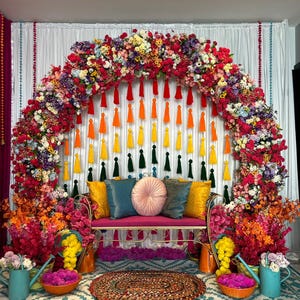 May include: A colourful, festive scene featuring a floral archway and a bench with cushions. The arch is decorated with flowers and tassels. A patterned rug and decorative elements complete the celebratory setting, creating a joyful atmosphere.
