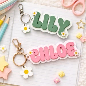 May include: Two custom keychains featuring the names LILY in dark green and CHLOE in pink, both with white backing and small daisy accents. Each keychain includes a gold-toned metal lobster clasp, a circular ring, and a white daisy charm.