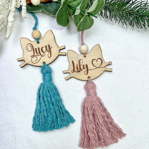 May include: Two wooden cat-shaped ornaments with the names "Lucy" and "Lily" engraved on them. Each ornament has a tassel, one is teal and the other is pink.