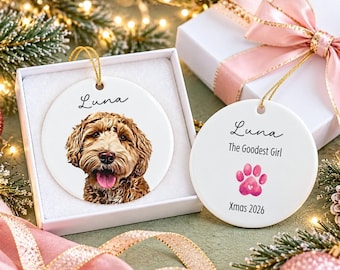 May include: Two round white ceramic ornaments with gold hanging strings. One ornament features a portrait of a brown dog with the name Luna. The other displays the name Luna, the text The Goodest Girl, a pink paw print, and Xmas 2026.