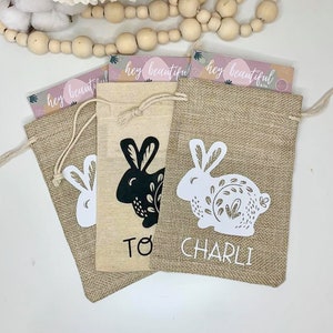 May include: Three burlap gift bags with bunny rabbit designs and the text "hey beautiful" and personalized names "To", "Charli".