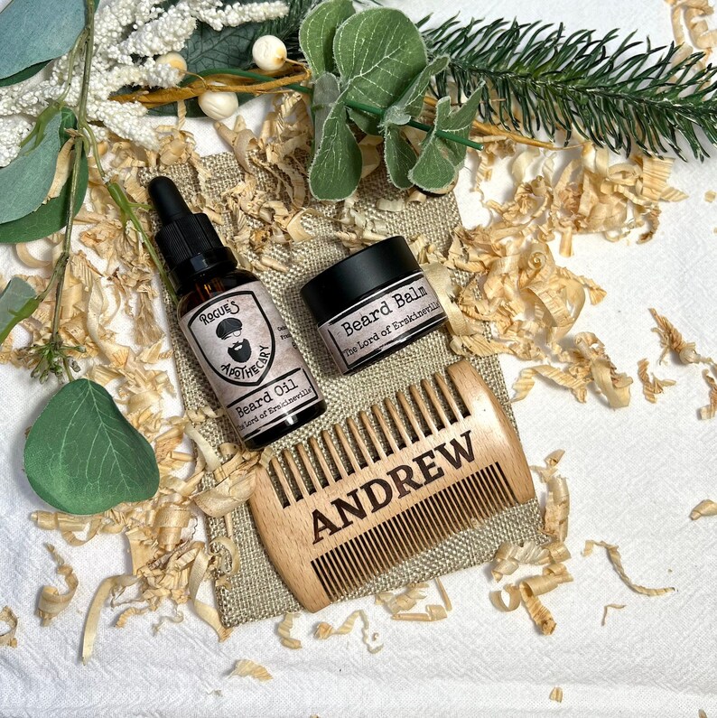 K&ouml;nnte beinhalten: Ein Holzkamm mit dem eingravierten Namen "ANDREW", ein kleines Glas mit Bartbalsam mit der Aufschrift "Beard Balm The Lord of Brackenhill" und eine Flasche Bart&ouml;l mit der Aufschrift "Rogue's Apothecary Beard Oil The Lord of Brackenhill" auf einer Jutestoff-Oberfl&auml;che.