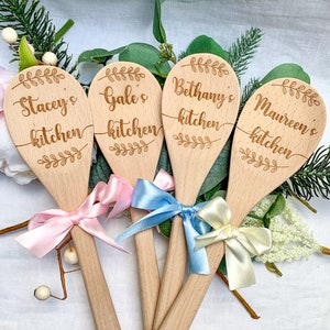 May include: Four wooden spoons with personalised text, each with a different name and the words "kitchen" engraved on them. The spoons are tied with ribbons in pink, blue and cream.
