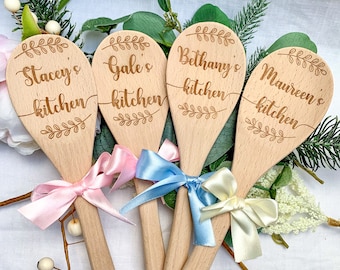 May include: Four wooden spoons with personalized text, each with a different name and the words "kitchen" engraved on them. The spoons are tied with ribbons in pink, blue, and cream.