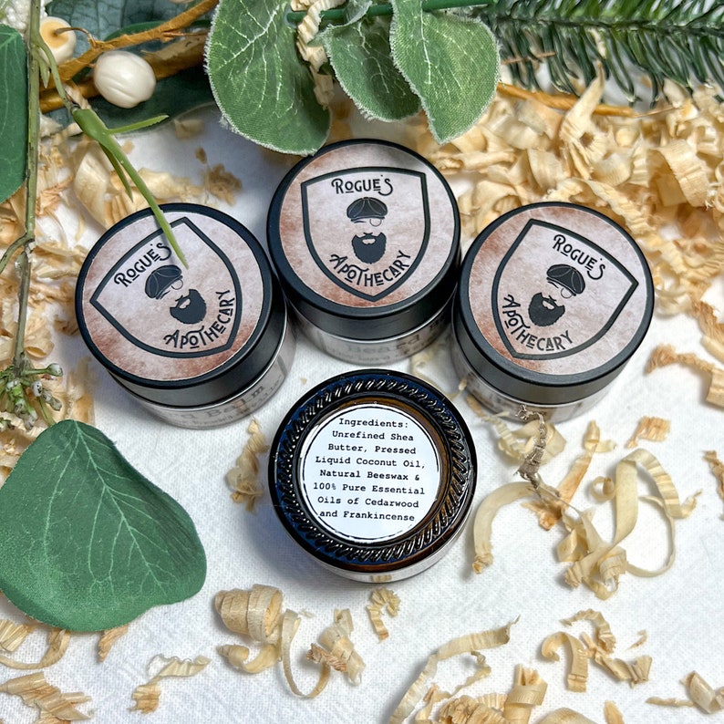 K&ouml;nnte beinhalten: Drei kleine Glasgef&auml;&szlig;e mit schwarzen Deckeln und Etiketten, auf denen "Rogue's Apothecary" steht. Die Etiketten zeigen eine Cartoon-Illustration eines Mannes mit Bart und Hut. Die Gef&auml;&szlig;e stehen auf einer wei&szlig;en Oberfl&auml;che mit Holzsp&auml;nen. Das Etikett auf einem Gef&auml;&szlig; listet die Zutaten auf: Unraffinierte Sheabutter, gepresste Kokosnuss&ouml;l, nat&uuml;rliches Bienenwachs und 100% reine &auml;therische &Ouml;le aus Zedernholz und Weihrauch.
